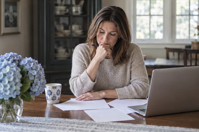 A Massachusetts homeowner reviewing an unmarked home-sale planning sheet beside a laptop at the dining table.